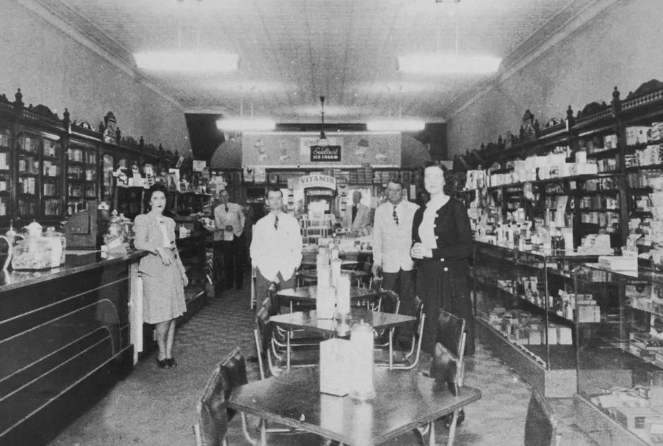 A vintage black-and-white photograph of the Gray Drug Company interior, with staff lined up between long wood shelving stocked with merchandise, soda fountain tables in the foreground, and an ice-cream sign at the rear.