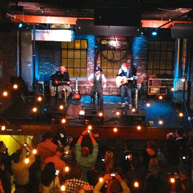 A view from the third-floor balcony looking down at the second-floor stage during a show — three musicians performing under stage lights with the crowd in the foreground.