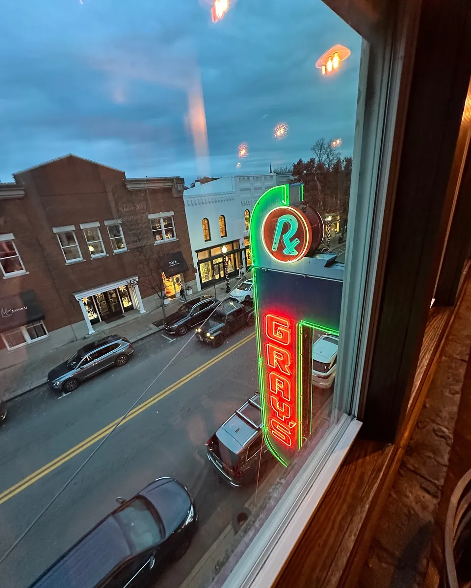 A view through a third-floor window at dusk showing the glowing red Gray's neon sign against the brick face and downtown Main Street below.